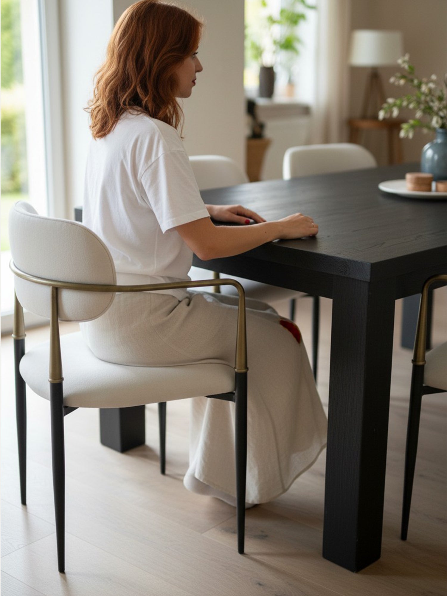 Elegance metal sandalye, açık renk döşemeli oturma ve sırt yapısıyla siyah metal ayaklar ve pirinç tonlu kollu tasarımıyla modern bir yemek masasında ev içi kullanımda yandan görünüm.
