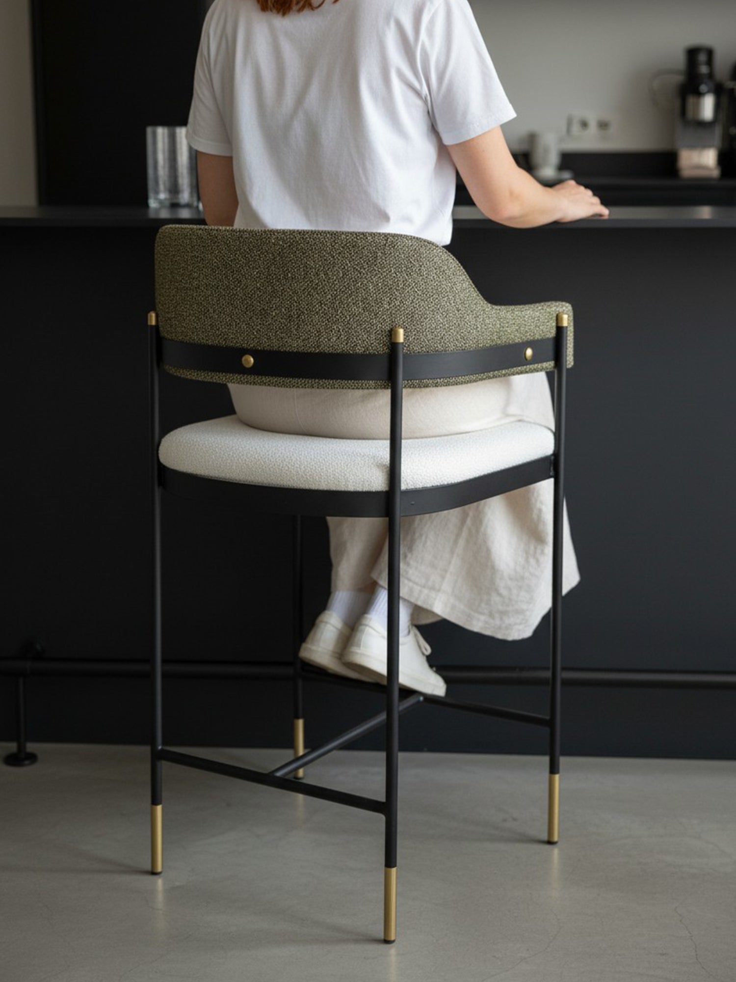 Siyah metal gövdeli, pirinç tonlu uç detaylarına sahip, gri dokulu döşemeli sırt ve kollu oturma tasarımı bulunan modern Elmor bar sandalye; iç mekânda bir kişinin oturduğu, bar tezgâhı yanında konumlandırılmış yaşam alanı çekimi.
